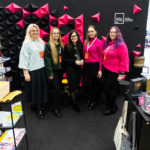 KTU leidyklos atstovės Vilniaus knygų mugėje KTU leidyklos atstovės prie KTU leidyklos stendo Vilniaus knygų mugėje. Jos stovi prieš juodą sieną su iškeltomis geometrinėmis juodomis ir ryškiai rožinėmis trikampių formomis, ant sienos matomas baltas „ktu“ leidyklos "technologija" logotipas.