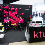 KTU leidyklos „Technologija“ stendas Vilniaus knygų mugėje Dvi KTU leidyklos atstovės sėdi prie KTU leidyklos stendo Vilniaus knygų mugėje. Jos sėdi prieš juodą sieną su iškeltomis geometrinėmis juodomis ir ryškiai rožinėmis trikampių formomis, ant sienos matomas baltas „ktu“ leidyklos "technologija" logotipas.