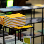 KTU leidyklos „Technologija“ knygos Vilniaus knygų mugėje Krūva geltonų kietais viršeliais knygų su derančiais geltonais juostiniais skirtukais ant juodo metalinio ekspozicinio stalo. Knygos tvarkingai sukrautos centre, o šalia jų padėtos kelios plonos geltonos skirtukų kortelės. Fone ant kitų juodų metalinių stalų matyti dar knygų krūvos, tačiau jos yra nefokusuotos.