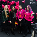 KTU leidyklos atstovės Vilniaus knygų mugėje KTU leidyklos atstovės prie KTU leidyklos stendo Vilniaus knygų mugėje. Jos stovi prieš juodą sieną su iškeltomis geometrinėmis juodomis ir ryškiai rožinėmis trikampių formomis, ant sienos matomas baltas „ktu“ leidyklos "technologija" logotipas.